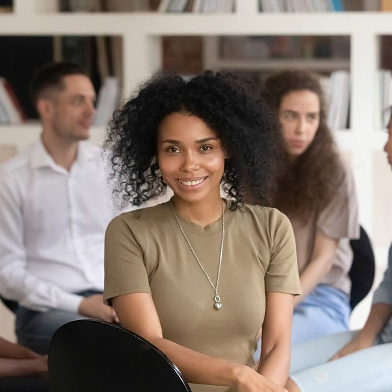 in a group of people sitting in chairs a woman turns towards the camera and smiles in an outpatient alcohol treatment program